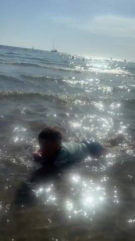 The video shows a young child lying on their stomach in shallow ocean water. The child is wearing a long-sleeved rash guard. The sun is shining brightly, creating a glare on the water. In the background, there are sailboats and other people in the water. The child appears to be looking towards the camera.