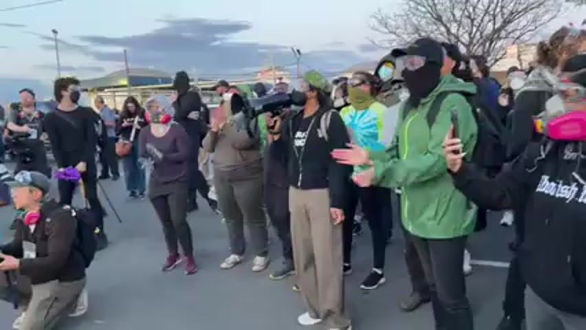 A group of approximately 15-20 individuals is gathered outdoors, appearing to be in a street or parking lot. Most participants are wearing face coverings such as balaclavas, gaiters, goggles, and respirators with pink, black, or grey filters.

In the center, one person holds a megaphone and leads a chant, saying "Close Grady Hall!" which the crowd repeats. This individual is wearing a black hoodie with partially visible text "BLK LIVES" and tan pants. To their right, another person in a green jacket and balaclava holds a smartphone, seemingly recording the event. To the far right, a person in a black "Abolish Ice" hoodie also wears a respirator. On the far left, a person is kneeling beside a tripod, possibly holding a camera.

The group is clapping and chanting in unison. Buildings and bare trees are visible in the background under an overcast sky, suggesting dusk or early morning. The ground is asphalt. The scene depicts a protest or demonstration.
