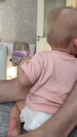 A toddler wearing a pink t-shirt and a diaper is seated on a couch with their back to the camera. A person's legs are visible to the right and left of the toddler, suggesting they are holding or sitting next to the child. The toddler's head is turned to the left. The background appears to be a room with patterned wallpaper and a dresser or cabinet. The lighting is soft and natural.