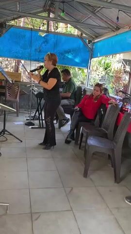 A blonde woman in black pants and a black top is singing into a microphone. She is standing to the left of a keyboard player, who is wearing a green shirt. A woman in a red top and black pants is sitting in a chair to the right of the keyboard player. A man in a blue shirt is standing behind the woman in the red top, facing away from the camera. There are chairs and tables in the background, and the scene appears to be outdoors under a covered patio. The time code indicates the video is 4 seconds long.