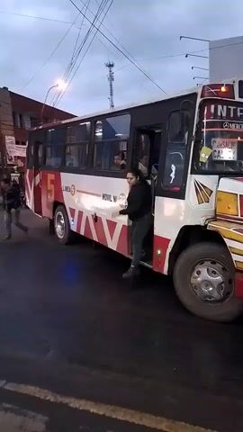 A woman exits a white and red bus parked on a wet street.  The bus has the number 5 and the words "LINEA 6" written on it.  A man walks past the bus.  The sky is overcast. Streetlights and wires are visible overhead.  Buildings line the street in the background.