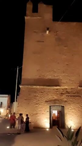 The video shows a nighttime scene in front of a tall, stone building, possibly a tower or castle, illuminated by spotlights at its base. Several people are standing and walking in front of the building. To the right, there appears to be an outdoor seating area for a restaurant or cafe with tables and people seated. The area is paved, and there are planters with greenery. In the foreground, parts of bicycles are visible. The sky is dark.