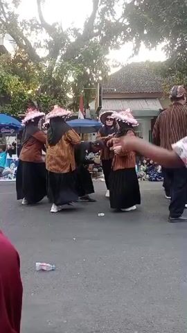 The video shows a street performance with several people in traditional clothing. The central group consists of at least five individuals wearing patterned shirts, dark skirts, and elaborate headdresses. They appear to be engaged in a synchronized dance, with some holding baskets. One person in the center is bent over. To the right, another individual in similar attire is also dancing. A man in a maroon shirt and black pants is walking from left to right in the foreground, holding a water bottle. In the background, there are spectators sitting and standing, with some trees and buildings visible. There is a small flag on a pole and what appears to be a tent or canopy on the left. The ground is paved asphalt, and there is a plastic bottle on the ground. The lighting suggests it is daytime.