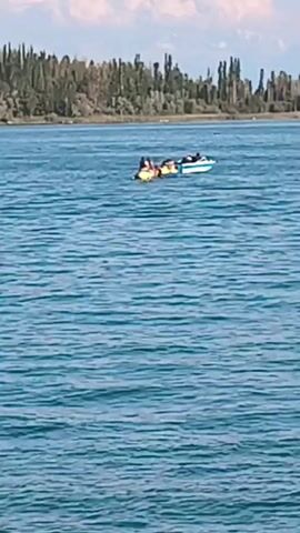 A motorboat tows an inflatable watercraft with several people aboard across a blue body of water. The watercraft appears to be yellow and rectangular. The motorboat is white with a blue stripe. In the distance, a treed shoreline is visible under a sky with scattered clouds.