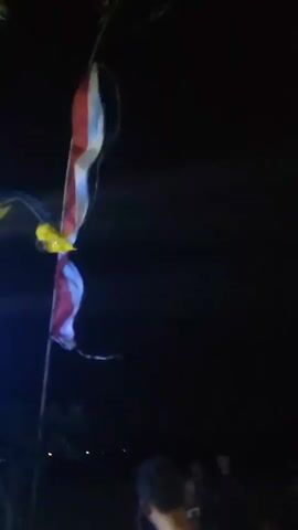 The video shows a group of people at night, with a sound system and lights in the background. The sound system is a large setup with many speakers and flashing lights. The lights are colored blue, green, and purple, and they are flashing in a pattern. There are several people in the video, some of whom are watching the sound system and lights. One person is walking towards the sound system, and another person is standing in front of it. The environment appears to be outdoors, with trees and foliage visible in the background. There is also a flag with red and white stripes hanging from a pole in the background. The video is shaky and the lighting is low, making it difficult to see details clearly.
