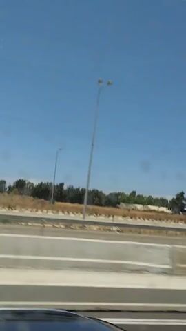 The video shows a view from a moving vehicle, likely on a highway. The sky is clear and blue. In the middle ground, there is a highway with multiple lanes and a barrier. Beyond the highway, there is a line of trees and some buildings, possibly industrial or agricultural. There are also tall structures that appear to be cell towers or light poles. Some vehicles are visible on the highway, including a white car that is blurred due to motion. The overall impression is a landscape typical of a developed area seen from a road.