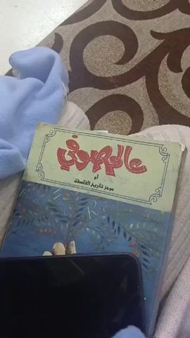 The video shows a close-up shot of a book titled "Sophie's World" in Arabic. The subtitle says "A Novel About the History of Philosophy." The book is on top of what appears to be a blanket or cushion with a light color. Part of a blue cloth is also visible. A black smartphone is partially obscuring the book. The background consists of a brown and beige patterned surface. The video is stable, with no camera movement.