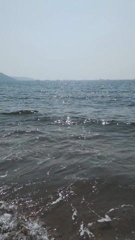 The video depicts a serene ocean scene with gentle waves lapping onto a dark sandy beach. In the distance, a hazy landmass is visible. The water is a deep blue with shimmering reflections of sunlight. The sky is light and clear, suggesting a bright, sunny day. There are no people or objects visible beyond the natural elements of the scene.