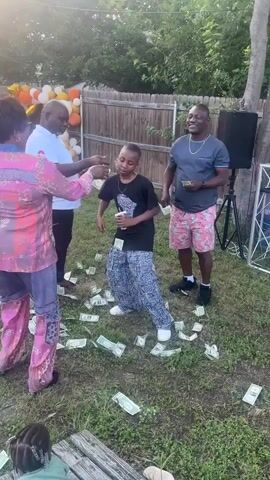 In a backyard setting, a boy in a black t-shirt and patterned pants dances on a lawn covered with scattered US dollar bills. He is surrounded by three adults who are either throwing money or looking on. A woman in a pink patterned outfit is partly visible to the left, and two men, one in a white shirt and one in a grey t-shirt and floral shorts, are standing to the side. A sound system is set up nearby, and a balloon display with orange and white balloons stands in the background near a wooden fence. A child is sitting on a wooden bench in the lower left corner. The event appears to be celebratory.