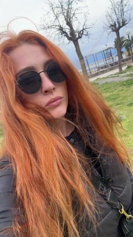 A woman with long orange-red hair is outdoors, standing on grass. She's wearing dark sunglasses and a dark jacket. The background shows trees, grass, a metal fence, and in the distance, water. The woman is facing the camera and speaking.