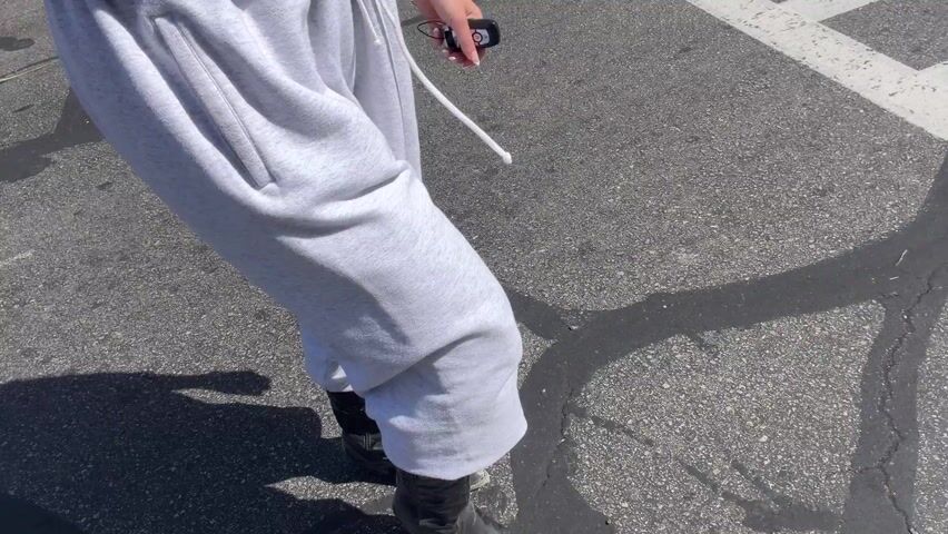 The video shows a person wearing gray sweatpants and black cowboy boots walking on asphalt. The person is holding a small black object in their right hand. The asphalt has white painted lines on it. The scene appears to be outdoors in daylight.