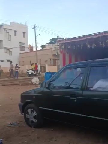 The video shows an outdoor scene in what appears to be a residential area, possibly in India, given the architecture and the presence of what looks like a tractor. The ground is dirt and gravel, suggesting it might be an unpaved street or lot. In the foreground, a blue car is parked, and a portable generator is visible near it.

In the middle ground, several people are gathered. Some individuals are wearing red and blue shirts, possibly uniforms, and are standing around a car and a sound system setup. A tractor is also visible. A person in a red shirt with a black pattern is standing and facing away from the camera, holding what appears to be a knife or a similar object. Nearby, a young person in a blue and white patterned outfit is standing. Several other people, including children, are standing around, some seemingly observing or participating in an activity.

In the background, there are multi-story buildings, consistent with a residential neighborhood. Utility poles and wires are present. The sky suggests it is daytime, possibly late afternoon or early evening, with some clouds visible.

On the right side of the frame, a white scooter is parked. Further down the street, more people are visible, some standing and others possibly moving.

In the latter part of the video, a vehicle, possibly an auto-rickshaw, is decorated with a large pink structure resembling a hand or a prop. A person is standing on top of this structure. In the immediate foreground, a drum is placed on the ground, and two people are seated nearby, one on a blue plastic chair. The overall impression is of a community gathering or event taking place.