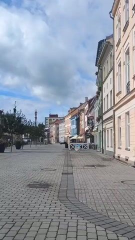 The video shows a cobblestone street lined with historic buildings in a European town. The buildings are of various heights and colors, with ornate architectural details such as balconies and decorative facades. Some of the buildings have shops or cafes on the ground floor, with awnings and outdoor seating visible. There are trees and potted plants along the street, and a fountain is visible in the distance. The sky is blue with some white clouds. A few people are seen in the distance, walking or sitting on benches, but they are not the focus of the video. The camera pans slowly, capturing the atmosphere of the street.