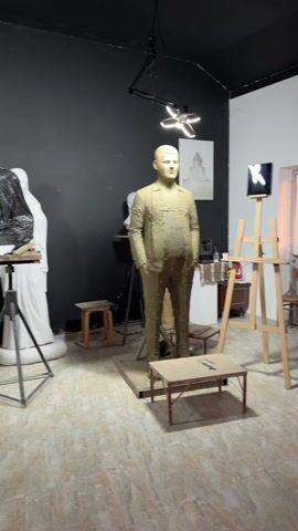 A man in a dark vest and blue jeans removes a black plastic covering from a sculpture in a studio. The studio has dark walls and a light floor. In the studio are various sculptures, stands, easels, and work surfaces. The room is lit by overhead lights.