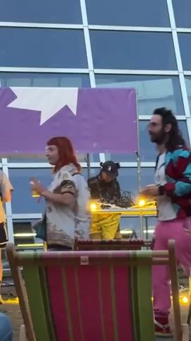 A group of people are gathered outdoors in front of a modern building with a glass facade and a large purple banner with a white star on it. In the foreground, two striped deck chairs are visible. In the mid-ground, a woman with red hair and a patterned shirt is holding a drink and talking. Next to her, a man with long dark hair, glasses, and a colorful jacket over a white t-shirt and pink pants is dancing. Behind them, a person wearing a black hat and yellow clothing is standing near DJ equipment. The sky appears to be dusk.