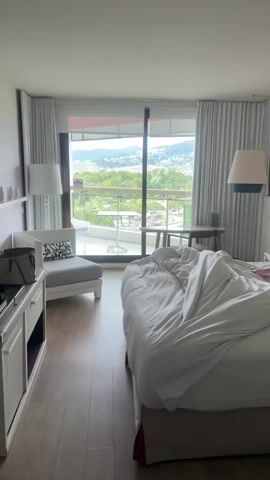 The video shows a bright hotel room. A large, unmade bed dominates the foreground.  Behind the bed is a large sliding glass door leading to a balcony. The balcony has a small table, chairs, and a partial view of the water and a mountainous landscape in the distance.  To the left of the sliding glass door is a grey armchair with a black and white patterned pillow and a white cabinet.  Two lamps, one on either side of the bed, provide additional lighting. The floor is wood.  The curtains are white.