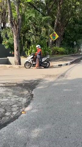 A person wearing a helmet and an orange hijab is standing next to a white scooter on the side of a paved road. The person is looking down at their phone. The scooter is parked on the left side of the frame, facing away from the camera. In the background, there are trees, foliage, and a yellow traffic sign with a red and green light symbol. The road is asphalt and appears to be in a suburban or semi-urban area. The lighting suggests it is daytime.