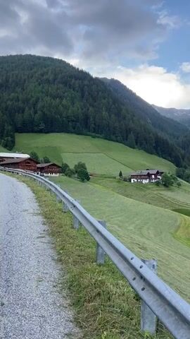 This media file is a vertical video showcasing a scenic mountain landscape in Ratschings - Racines, Italy. The video opens with a view of a paved road curving to the left, bordered by a metal guardrail and a grassy slope. Beyond the road, traditional alpine houses with dark roofs are nestled on green, mowed hillsides.

The perspective then shifts to reveal the grandeur of the surrounding mountains. A prominent, densely forested mountain dominates the frame, its slopes covered in a thick carpet of evergreen trees. As the video progresses, the view expands to encompass a valley, with more tree-covered mountains receding into the distance. In the valley, a small village with white buildings is visible, set against a backdrop of lush green fields.

The time of day appears to be late afternoon or early evening, with the sky filled with dramatic, cloudy formations. Sunlight filters through the clouds in some areas, casting a soft glow on the peaks. The weather seems to be transitioning, with a sense of impending change suggested by the cloud cover.

The overall mood conveyed is one of tranquility and natural beauty. The vastness of the mountains, the charming architecture of the houses, and the serene valley evoke a sense of peace and awe. There is no visible text within the media file.
