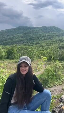 The video shows a woman with long brown hair wearing a dark jacket, blue jeans, and a gray baseball cap. She is sitting on a rocky outcrop overlooking a lush green valley. Behind her, a large, tree-covered mountain rises under a cloudy, overcast sky. The camera slowly pans upward, shifting the focus from the woman to the landscape, ultimately centering on the dramatic sky and mountain view.