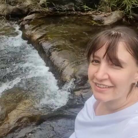 A woman with brown hair pulled back into a bun with a side fringe is seen in a selfie-style video. She is wearing a white t-shirt and is standing next to a small waterfall with rocky edges and surrounding greenery. The woman smiles at the camera and points towards the waterfall with her right hand.