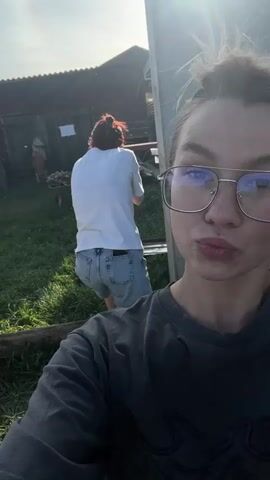 The video shows two women outside on a sunny day, possibly in a rural area. One woman, with glasses and her hair in a bun, is holding the camera, taking a selfie-style video. Behind her, another woman with reddish hair pulled back is climbing a makeshift ladder leaning against a large, gray metal structure. The ladder appears to be made of wooden planks attached to a metal frame. The woman climbing is wearing a white t-shirt and jean shorts. In the background, there's a wooden building, possibly a shed or small house, and a wheelbarrow with wood in it. The area has grass and a few scattered objects. The sun is bright, creating a lens flare in the video. The camerawoman briefly stops moving the camera, revealing a third person with long dark hair to the left of the woman climbing the ladder.