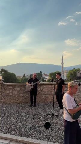 A man plays an acoustic guitar and sings into a microphone while another man stands near him, also singing. They are on a stone patio with a stone wall behind them. In the background, there are buildings and mountains. The sky is partly cloudy.