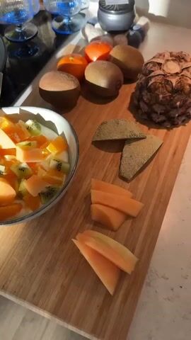 The image captures a bright, domestic kitchen scene in Bergen, Norway, bathed in warm, directional sunlight, likely during the morning or late afternoon. A wooden cutting board dominates the right side of the frame, laden with fresh fruit. On the board are a whole pineapple with its characteristic spiky texture, three fuzzy brown kiwi fruits, and two vibrant orange mandarins. Partially peeled and sliced cantaloupe melon is also displayed, with two large pieces showing their netted green skin and several smaller, peeled, orange wedges arranged below them.

In the foreground, to the left, a white ceramic bowl with a dark rim contains a creamy white base, possibly yogurt, topped generously with diced orange cantaloupe, bright green kiwi pieces, and segments of orange. A silver spoon rests in the bowl. Behind the bowl, on a black stovetop, a round, light brown pancake or crepe is cooking in a frying pan.

The background reveals further kitchen details: a white pot, two blue-tinted, ribbed glass tumblers, and two ingredient bags. Visible text on one bag reads "CELTIC HAVSSALT" (Celtic Sea Salt), and the other partially shows "OPULVER". The overall scene suggests a healthy breakfast or snack being prepared in a well-lit, inviting space.