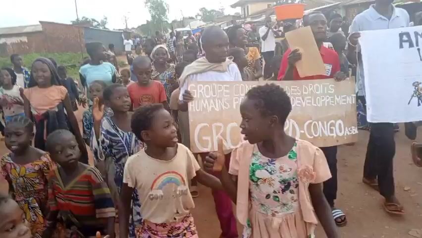 A group of adults and children are marching down a red dirt road through a village. They are carrying signs written in both French and what appears to be a local language. The signs seem to be related to human rights and development in the Congo. The people are mostly dressed in casual clothing. Some buildings can be seen in the background. A police officer in uniform is also present in the crowd. The atmosphere appears to be one of determined protest.