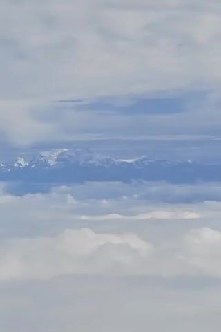 A video showing a wide view of snow-capped mountains peaking above a layer of clouds.