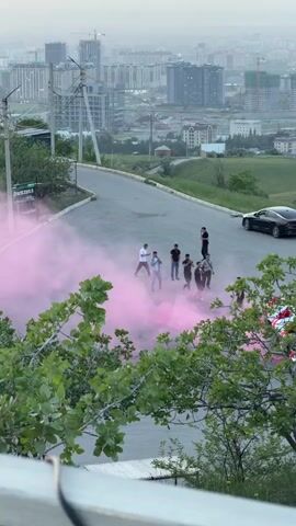 A group of approximately 10 young men stand amidst a cloud of pink smoke on a sloping asphalt road. A black sports car is parked nearby. The background reveals a cityscape under a hazy sky, with buildings and construction cranes visible. Green trees frame the lower part of the view. The scene suggests a celebration or a gender reveal event.