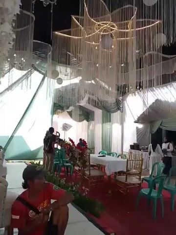 The video depicts an outdoor wedding reception. The space is decorated with white draping, dangling white ornaments, and red carpeting. Several tables and chairs are arranged for guests. People are seated, some wearing hijabs, while others stand and move around. Some individuals appear to be serving food. A few children are visible among the guests. The overall atmosphere is festive.
