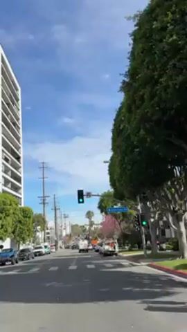The camera moves along a street in a city neighborhood, likely in the United States. The sky is blue with scattered white clouds.  A tall, modern building with horizontal balconies sits on the left side of the street. Utility poles line the road, and there are cars parked along both sides. Several utility or construction vehicles are visible further down the street.  A tree with pink blossoms is on the right. The traffic light shows a green light. Street signs are visible, one reading "Alta Loma Rd".