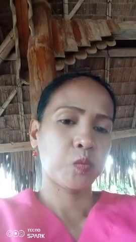 The video shows a close-up of a woman wearing a pink top. She has her hair pulled back and is wearing earrings. The background appears to be a thatched roof structure with wooden beams. The woman makes a puckering motion with her lips and then stops. The video is short and appears to be a selfie.