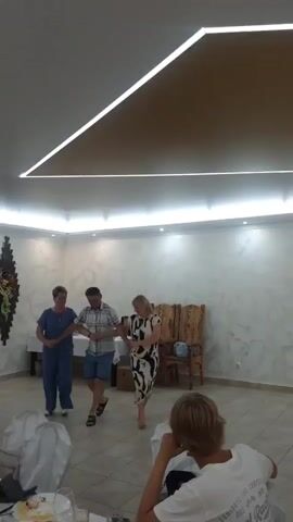 Three people are dancing in a room. The person on the left is a woman wearing blue scrubs. The person in the middle is a man wearing a plaid shirt and blue shorts. The person on the right is a woman wearing a black and white patterned dress. The three individuals are holding hands and appear to be performing a coordinated dance. They are in a room with beige tiled flooring and white textured walls. There are chairs in the background, and a table with items on it. The ceiling has modern recessed lighting, with one prominent diagonal light fixture. The scene appears to be indoors, possibly at a party or event. There are two children in the foreground, facing away from the camera and watching the dancers. One child is wearing a white t-shirt with text on it. The text on the t-shirt is partially visible and appears to include "COMPETITION" and "1978". The lighting suggests it is daytime or well-lit indoors. The overall atmosphere seems festive and joyful.