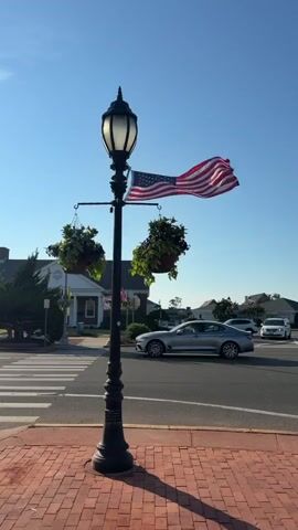 A lone lamppost stands on a brick sidewalk, adorned with an American flag and two hanging flower baskets. The flag is waving in the wind. Behind the lamppost, a crosswalk is visible on a street with a few cars driving by. The sky is clear and blue, suggesting a bright, sunny day. The overall scene depicts a typical streetscape, possibly in a small town or suburban area.