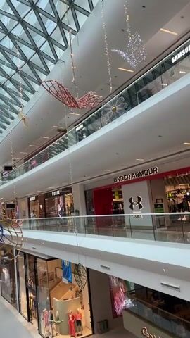 This is a vertical, eye-level, full shot video of a large, modern, multi-level shopping mall. The camera is positioned to look down an open atrium with a curved, glass ceiling that allows natural light to filter in. Hanging decorations, including large, stylized butterflies made of wire and lights, are suspended throughout the atrium. Shops are visible on each level, with store names like Guess and Under Armour clearly discernible. People are seen walking on the ground level and on the elevated walkways. The environment appears clean and well-lit, with a sense of scale and openness.
