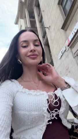 A woman with long dark hair and makeup is taking a selfie outdoors. She is wearing a white cropped sweater with bell sleeves and a brown top underneath, paired with blue jeans. She is also wearing a necklace with a cross pendant and large hoop earrings. The background appears to be a city street with buildings.