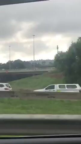 The video shows a view from inside a moving car, looking out the side window. Outside, there is green grass and a concrete highway overpass. Several light poles line the side of the highway. There are cars on the overpass. The sky is overcast and gray.