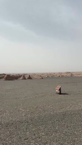 The video displays a vast, desolate, and arid landscape under a hazy, overcast sky. The foreground is a flat expanse of dark gravel. A small, dark brown, house-like wooden structure stands isolated in the center. In the mid-ground, a long band of eroded rock formations, typical of a yardang landform, stretches across the horizon, appearing somewhat indistinct due to the atmospheric haze.

At 0:02, the camera pans slightly to the right, revealing an asphalt road running straight into the distance on the right side of the frame, marked by a solid white line. A person wearing a dark jacket, dark pants, and a wide-brimmed hat is walking away from the camera along the side of the road. The distant rock formations remain visible. The scene suggests a remote, desert-like environment, consistent with locations around Dunhuang City, China. No visible text appears in the video.