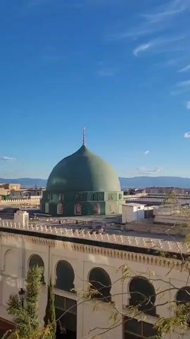 The video pans slowly across the rooftops of a building complex, culminating in a shot of the sun shining against a clear blue sky. The camera starts focused on the green dome of a mosque or similar building, complete with a crescent moon topper. The complex features cream-colored walls with repeating arch patterns. Beyond the buildings, a cityscape can be seen with mountains on the horizon. As the camera pans right, the sun comes into view, bright and partially obscured by atmospheric haze. There are a few wispy clouds in the otherwise clear sky. A bare tree branch is visible in the foreground throughout.