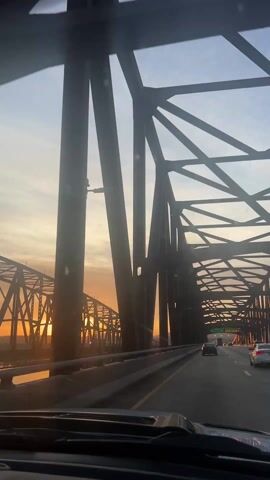 A car drives on a bridge as the sun sets. The bridge is made of metal beams. Other cars are on the bridge. The sky is orange and blue. The camera is inside the car.
