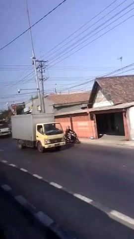 The video shows a street scene from a moving vehicle. In the foreground, there is a road with white dashed lines. In the background, there are buildings, trees, and power lines. Several vehicles are visible, including a yellow box truck, a silver van with red and orange graphics, a white truck with a flatbed, a white SUV, and a few motorcycles. People are seen on the motorcycles and possibly in the vehicles. The sky is blue and clear. There are also some Indonesian flags visible on poles. The lighting suggests it is daytime.