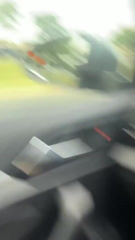 The video shows a white Toyota Supra driving on a road. The car is driving in the opposite direction to the camera. The car has black wheels and red brake calipers. The car has a spoiler and a blue exhaust tip. The video is taken from inside a car. The trees and street lights can be seen in the background.