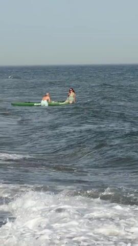 Two people are on paddleboards in the ocean. One person is sitting on a green paddleboard and the other is sitting on a white paddleboard. The person on the green paddleboard is wearing blue shorts and the person on the white paddleboard is wearing a light-colored swimsuit. The waves are rolling in and there is white foam on the water. The sky is clear and blue.