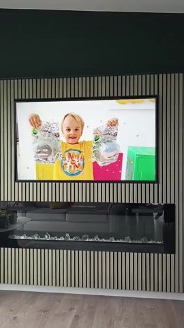 A television screen mounted on a slatted wall displays a video of two people, an adult woman and a young child, engaged in an activity. The woman, wearing a pink shirt and headband, is on the left side of the screen. The child, wearing a yellow shirt, is on the right. They appear to be pulling on a toy or object that is being stretched between them. The fireplace below the television is not actively burning, but has decorative glass pieces within it, and a sofa is visible behind it. The room has wooden flooring.