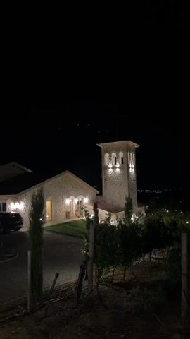 This is a nighttime video of a stone building complex with an attached tower, likely a winery or estate, set against a dark sky. The building and tower are illuminated by warm, ambient lights, highlighting the architecture. In the foreground, there are visible vineyard trellises and some dark foliage, suggesting the location is in a rural or semi-rural setting. A dark-colored vehicle is parked to the left of the building. The overall atmosphere is serene and somewhat elegant. There are no people or discernible actions captured in the video.