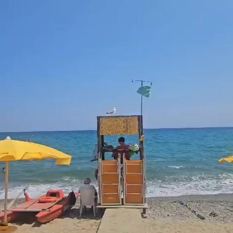 The video shows a beach scene on a clear, sunny day with a bright blue sky. In the foreground, a lifeguard stand made of wood and metal is positioned on the sand, facing the ocean. A seagull is perched on top of the lifeguard stand. Inside the stand, a person is sitting and appears to be looking down. In the background, the blue ocean stretches out to the horizon, with gentle waves breaking on the shore. A yellow umbrella is visible on the left side of the frame, and another person is partially visible sitting on the beach in the lower left corner. A green flag is flying from a pole attached to the lifeguard stand.