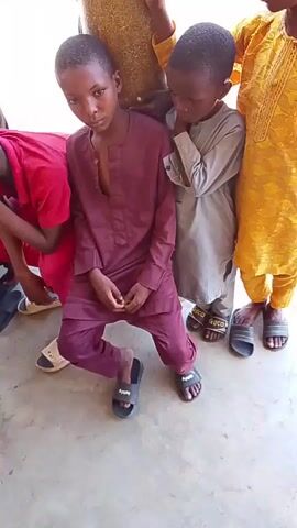 A group of four young boys stand near a cylindrical pillar. The boy closest to the camera, wearing a burgundy outfit, is seated on the ground with his legs bent. The other three boys are standing behind him. The boy to his right wears a light gray outfit. The boy on the far right wears a yellow outfit. To the left of the seated boy, another boy in a red shirt is partially visible. All the boys are wearing sandals. The floor appears to be concrete. The lighting suggests an outdoor or well-lit indoor setting.