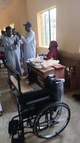 The video shows an indoor scene, possibly a classroom or community hall. In the foreground, a black wheelchair is parked. Behind it, a wooden desk is cluttered with various items, including a roll of white material, a bottle of hand sanitizer, and other objects that are difficult to identify. A woman wearing a red headscarf and a red and black patterned garment sits behind the desk.

To the left of the desk, three men are visible. One man, wearing a light blue traditional outfit and a dark cap, is looking at something in his hand. Another man, in a grey traditional outfit, stands next to him. A third man, wearing a white traditional outfit and a cap, appears to be interacting with the man in the blue outfit. Two wooden benches are also present in the scene.

Further in the background, more people are gathered, some of whom are wearing traditional attire. Sunlight streams in from a window on the right side of the room, illuminating dust motes in the air. The overall impression is of a gathering or event, possibly related to medical assistance or community support, given the presence of the wheelchair and medical supplies.