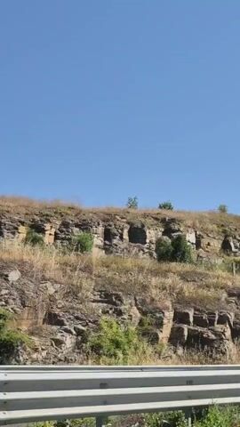 A video shows a clear blue sky above a rocky hillside covered with dry grass and scattered trees. A white barrier is visible in the foreground. As the camera pans to the right, a flagpole with a waving flag comes into view on top of the hill. The flag appears to have horizontal stripes of red, white, and green.